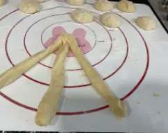 Step <p>Coloque la masa en una superficie de trabajo limpia y seca y ligeramente enharinada y divida la masa en 12 bolitas de aproximadamente el mismo tamaño. Coja 3 bolitas y forme unos cilindros con cada una de ellas de aproximadamente 50 cm de largo y junte los tres extremos y vaya formando una trenza, para finalizar vuelva a juntar los extremos inferiores para sellarlos y dele con la mano la forma de gaiato ( garrote)</p> illustration